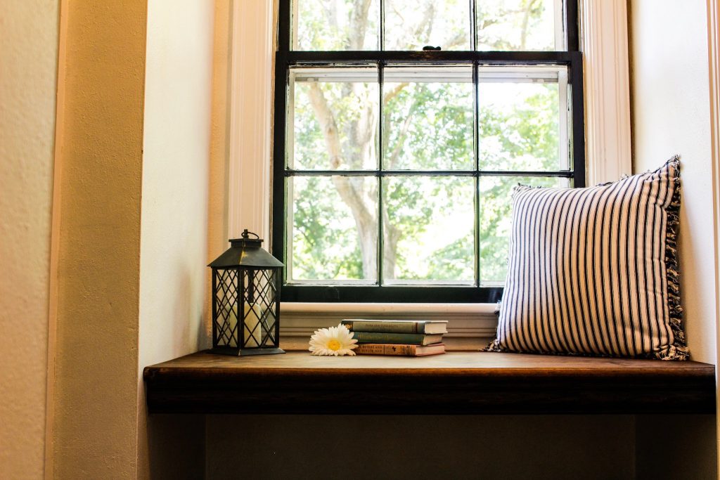 Walnut stain adds depth of color to the new window seat, which is surrounded by white walls and flanked by a paned window looking out at a huge, leafy tree.