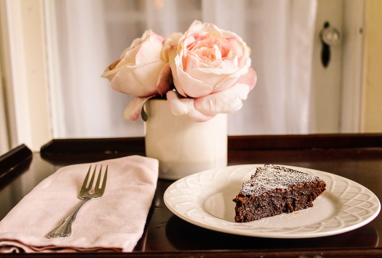 A piece of flourless chocolate cake sits on a white plate next to a vase of pink flowers and a pink napkin with a fork lying on it.