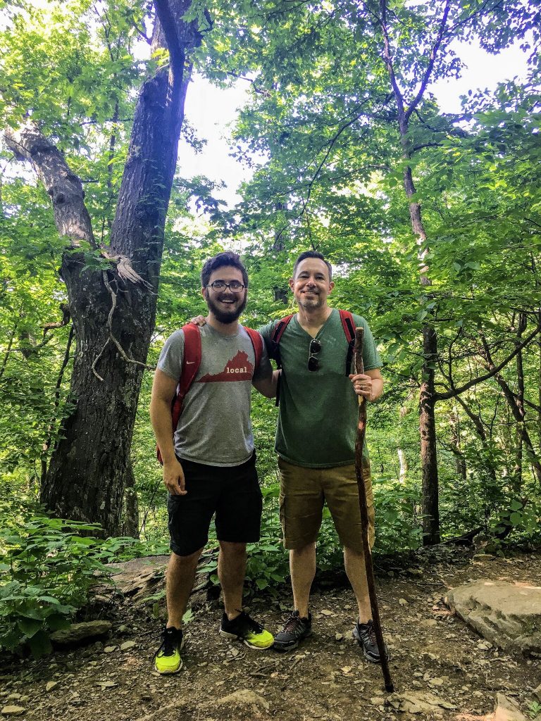 two men with backpacks in the woods-- gifts suggested for this guy are things such as an insulated backpack, down jacket, or paracord bracelet