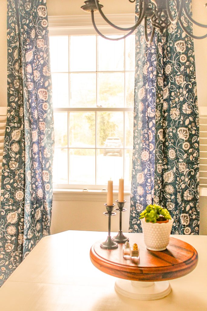 long, blue and white floral curtains framing open window; wood cake stand in foreground on top of antique white dining table, all part of our budget makeover
