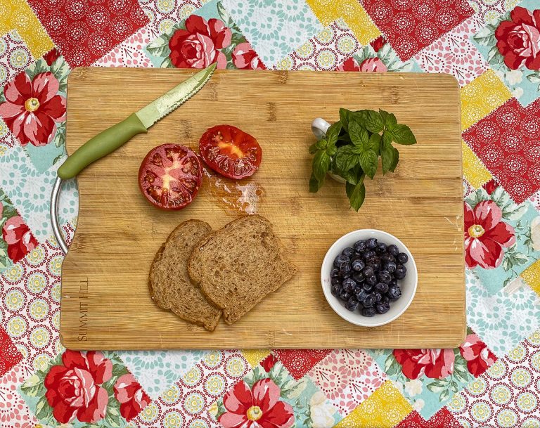 a wooden cutting board sits on a decorative tablecloth. On the cutting board are a sliced tomato, knife, slices of bread, blueberries, and a bunch of fresh basil.