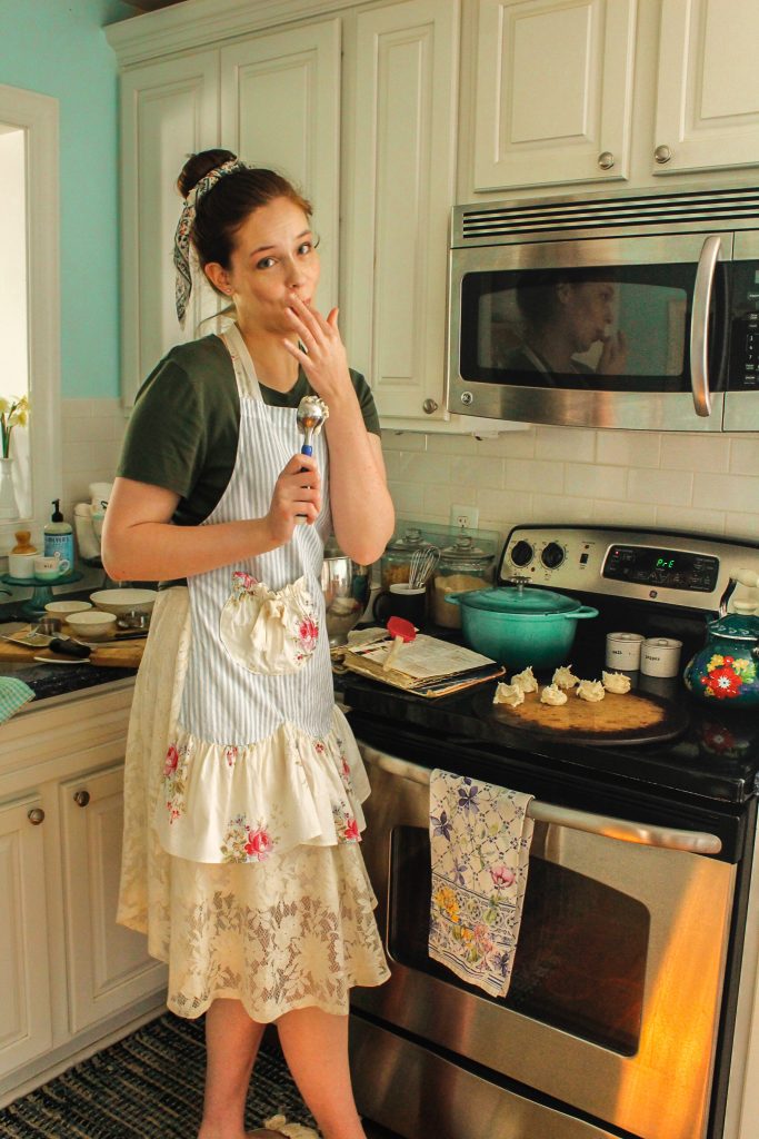 A young woman licks her fingers as she bakes cookies in a cute apron.